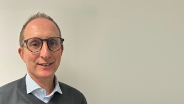 Professional headshot of a middle-aged man with glasses, wearing a dark sweater over a light blue striped shirt, smiling at camera against neutral beige background