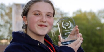 Young person holding a glass award trophy outdoors, smiling at camera, wearing navy hoodie with red lanyard