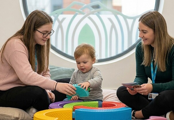 Two Ormiston staff members playing with a toddler using colourful stacking toys in a bright early years setting