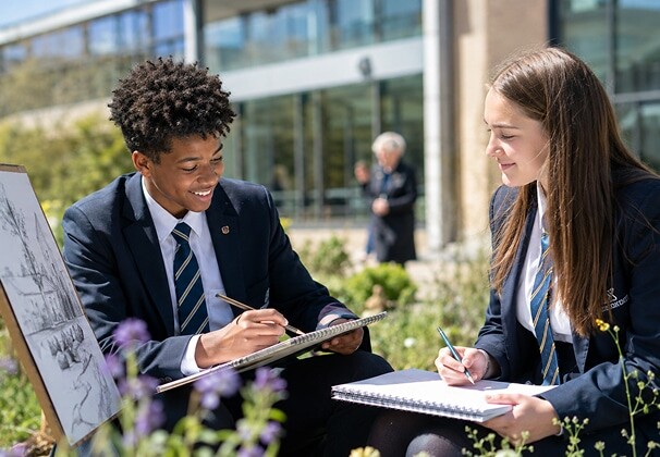 Two secondary school students sketching and writing notes outdoors on a sunny day