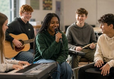 Female student singing into a microphone with peers playing guitar, keyboard and drums in a music room